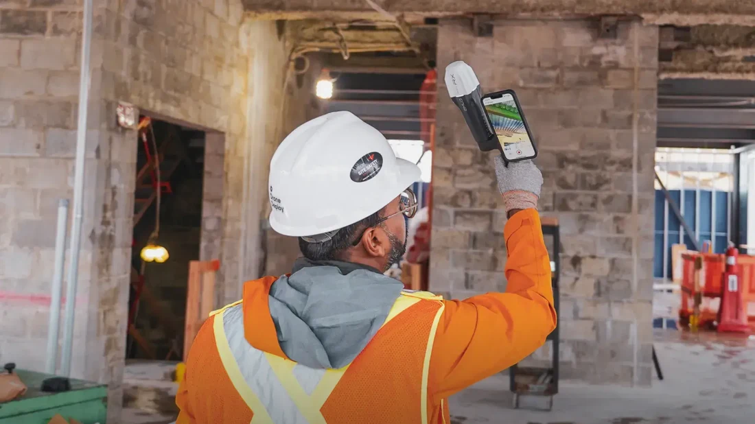 A construction contractor wearing a white DroneDeploy-branded hard hat and orange high-visibility safety jacket holds up a phone-mounted 360-degree camera capture device while standing inside a commercial concrete-block building under construction, illustrating the DroneDeploy Ground interior 360-walk capture workflow with native Procore data sync