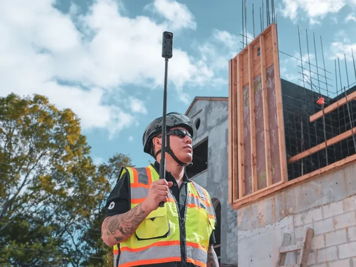 A construction worker in a high-visibility safety vest and dark hard hat holds an Insta360 360-degree camera mounted on a tall monopod above his head, with concrete forms, scaffolding and a partially-framed commercial building visible in the background, illustrating the DroneDeploy Ground capture workflow