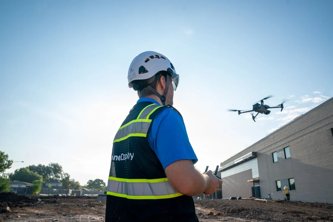 A construction contractor wearing a DroneDeploy-branded high-visibility vest and white hard hat operates a DJI drone over an active commercial construction site, with the drone visible in the upper right corner of the frame against a clear sky and a partially-completed building visible in the background