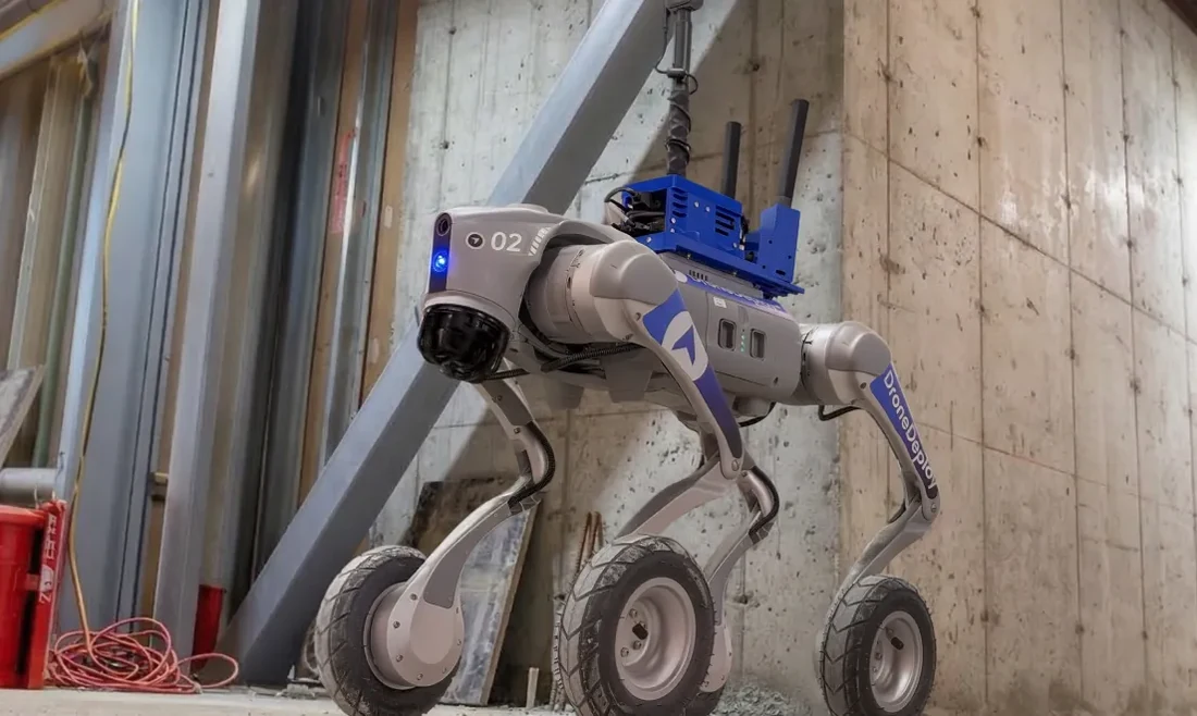 A DroneDeploy-branded autonomous ground robot with wheeled legs and a blue and white body labeled DroneDeploy stands inside a commercial construction site between concrete walls, with framing materials, electrical conduit and an exposed structural beam visible in the surroundings — representing the agentic-AI ground-robot product DroneDeploy announced for 2026 beta deployments